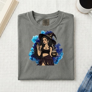 May include: A charcoal gray t-shirt featuring a graphic of a long-haired person wearing a witch hat, holding a smartphone and a cauldron. The design is set against a vibrant blue smoke background, with the Comfort Colors brand label visible.