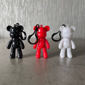 May include: Three small bear-shaped keychains in black, red, and white. Each bear has a glossy finish and a black clip for attaching to keys or bags. The keychains are standing upright on a gray surface.