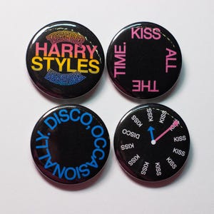 May include: Four round black pinback buttons with text in various colors. One button says "HARRY STYLES" in yellow and pink. Another says "KISS ALL THE TIME" in pink. The third says "DISCO, OCCASIONALLY" in blue. The last says "KISS" around a clock.