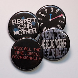 May include: Four black round buttons with white and red text. One button says "Respect Your Mother" with a globe graphic. Another says "Kiss All The Time. Disco, Occasionally". The other two say "We Belong Together" and "Kiss" around a clock graphic.