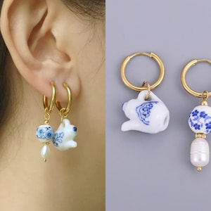 Puede incluir: Pendientes de aro dorados con abalorios de porcelana blanca y azul. Un pendiente presenta una pequeña tetera, el otro una cuenta azul y blanca con una perla colgante. Los pendientes se muestran sobre un fondo gris.