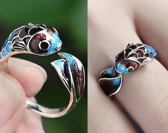 May include: A silver-toned ring featuring a detailed fish design. The fish is painted with blue, black, and maroon enamel, with the tail and head forming the ring's band. The ring is open, allowing for size adjustment. The ring is a unique piece of jewelry.