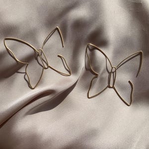 Puede incluir: Un par de pendientes de alambre en tono dorado con forma de mariposa. Cada pendiente presenta un contorno metálico fino y continuo que forma las alas y el cuerpo de una mariposa, creando un diseño delicado de marco abierto con un acabado pulido.