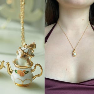 May include: A gold-colored necklace with a pendant featuring a white teapot with gold trim and a small animal inside. The teapot has orange floral accents. The necklace is displayed on a person wearing a burgundy top.