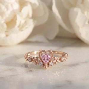 Pode incluir: Um anel de ouro rosa com uma pedra preciosa rosa em forma de coração no centro. O anel apresenta um design floral com pequenas pedras preciosas brancas e é colocado sobre um fundo desfocado de flores brancas.