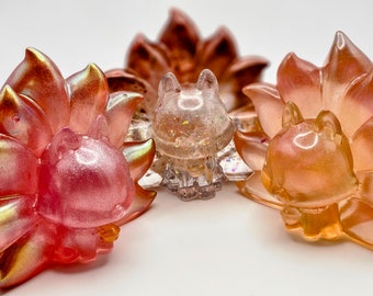 May include: Three small resin figurines of foxes with iridescent pink, orange, and clear finishes. The foxes have a stylized, cartoonish appearance with large ears and bushy tails.
