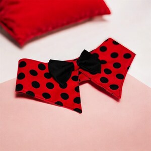 May include: A red and black polka dot collar with a black bow. The collar is red with black spots and a black bow tie. A red cushion is in the background.
