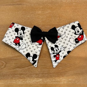 May include: A white fabric collar with a black bow tie and a Mickey Mouse print. The collar has a scalloped edge and features Mickey Mouse in various poses.