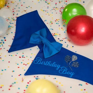 May include: A royal blue birthday sash with a matching bow tie. The sash reads "Birthday Boy" in script with a glittery balloon design. Colorful confetti and balloons in green, red, and white are scattered around the sash.