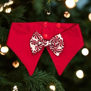 May include: A festive red pet collar with a bow tie. The collar has three red buttons and a bow tie with a white background and a red and burgundy swirl pattern. The collar is set against a background of a Christmas tree.