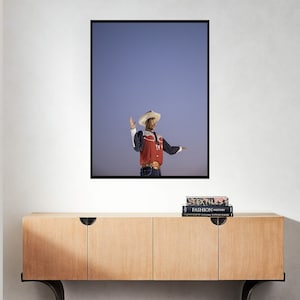 May include: A man wearing a cowboy hat and red shirt stands with his arm raised in front of a blue sky. He is wearing a belt with a large silver buckle. The image is framed and hanging on a white wall above a wooden cabinet.