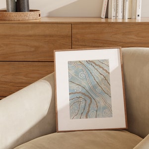May include: A framed artwork with a blue background and brown and white abstract patterns. The artwork is in a wooden frame and is sitting on a light brown couch.