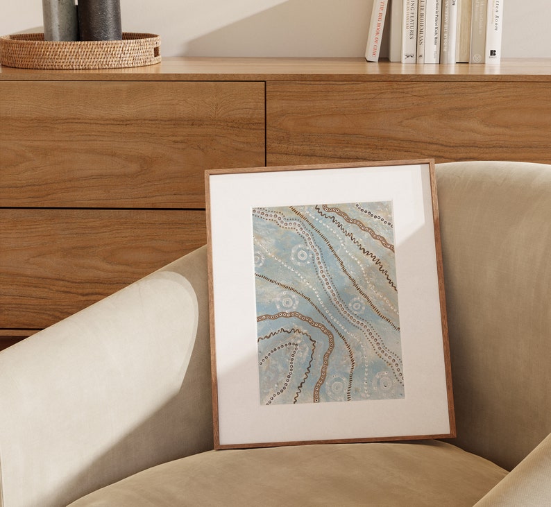 May include: A framed artwork with a blue background and brown and white abstract patterns. The artwork is in a wooden frame and is sitting on a light brown couch.