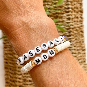 May include: Two white beaded bracelets with the words "BASEBALL" and "MOM" spelled out in black letters. The "MOM" bracelet has gold beads between the letters.