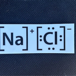 May include: A white sticker with black text and symbols showing the chemical formula for sodium chloride, [Na]+[Cl]-.