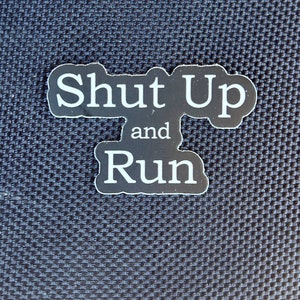 May include: Black and white sticker with the text "Shut Up and Run" on a dark blue textured background.