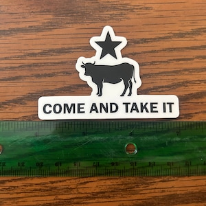 May include: A white sticker with a black silhouette of a cow and a star above it. The sticker says "COME AND TAKE IT" in black text.