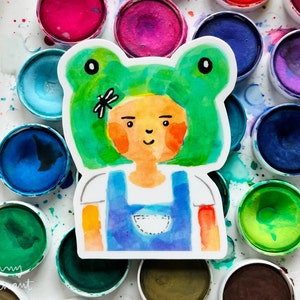 May include: A watercolour illustration of a person wearing a green frog hat with a dragonfly on top. The person is wearing a blue overall and has a white pocket on the front. The illustration is surrounded by watercolour paint circles.