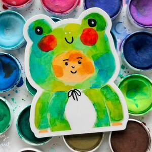 May include: A watercolour illustration of a frog wearing a green and blue frog costume. The frog is smiling and has a white face. The illustration is on a white background.