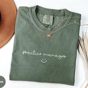 May include: A moss green t-shirt with the words "Practice manager" in white script and a smiling face. A gold necklace with a pendant is on the shirt. A brown hat and dried palm leaves are in the background.