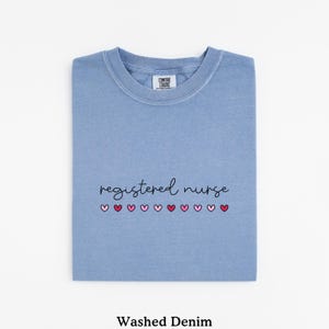 Pode incluir: Uma camiseta azul clara dobrada com as palavras "registered nurse" em escrita preta e uma fileira de cora&ccedil;&otilde;es rosa. A camiseta est&aacute; rotulada como "Washed Denim" na parte inferior.