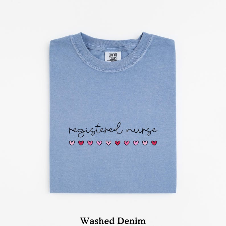 Pode incluir: Uma camiseta azul clara dobrada com as palavras "registered nurse" em escrita preta e uma fileira de cora&ccedil;&otilde;es rosa. A camiseta est&aacute; rotulada como "Washed Denim" na parte inferior.