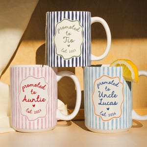 May include: Three ceramic mugs with vertical stripes in navy blue, pink, and light blue. Each mug features a central scalloped label with text: Promoted to Tio Est. 2027, Promoted to Auntie Est. 2027, and Promoted to Uncle Lucas Est. 2026.