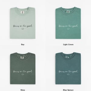 May include: Four folded t-shirts in different shades of green and blue. Each shirt has the phrase "focus on the good" and a smiley face printed on the front. The colors are Bay, Light Green, Moss, and Blue Spruce.