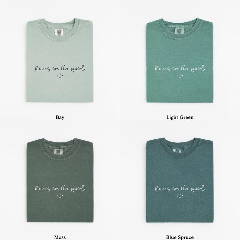 May include: Four folded t-shirts in different shades of green and blue. Each shirt has the phrase "focus on the good" and a smiley face printed on the front. The colors are Bay, Light Green, Moss, and Blue Spruce.