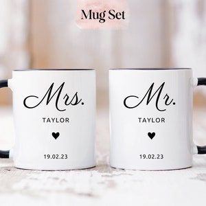 May include: A set of two white ceramic mugs with black handles and rims. The mugs have black text that reads "Mrs. TAYLOR" and "Mr. TAYLOR" with a small black heart and the date "19.02.23" below the text.