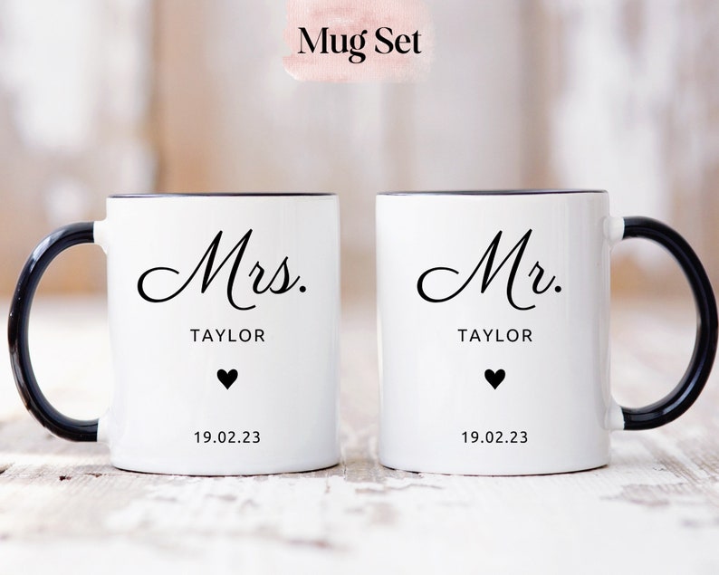 May include: A set of two white ceramic mugs with black handles and rims. The mugs have black text that reads "Mrs. TAYLOR" and "Mr. TAYLOR" with a small black heart and the date "19.02.23" below the text.
