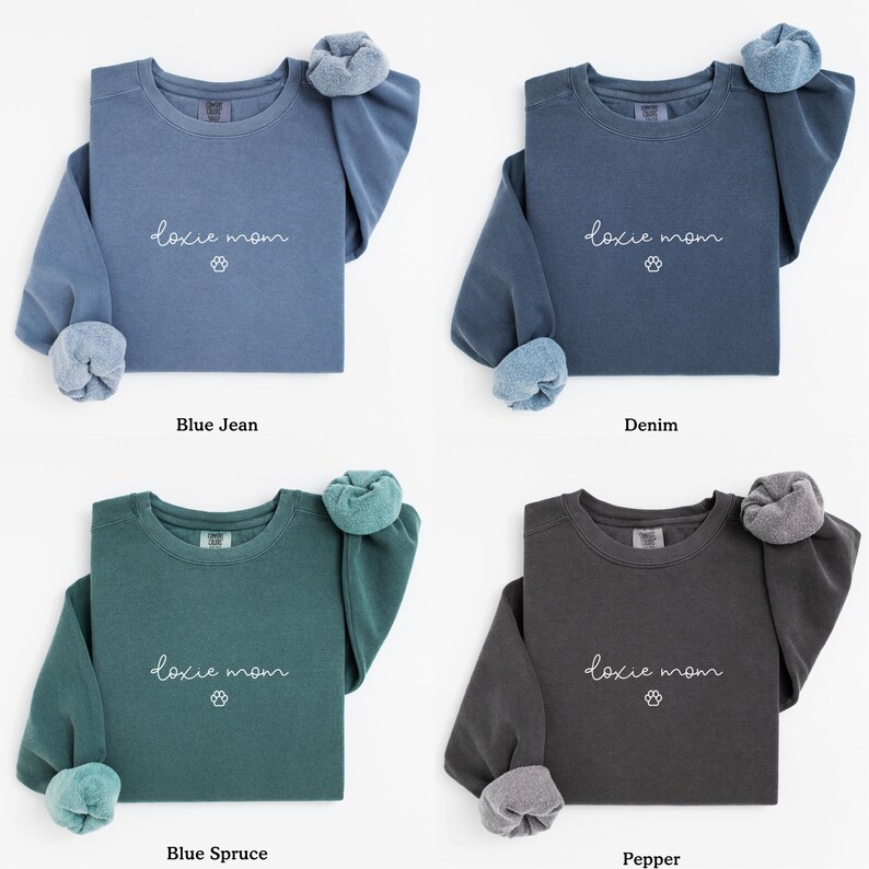 May include: Four sweatshirts in different colors: blue jean, denim, blue spruce, and pepper. Each sweatshirt has the text "doxie mom" in cursive with a paw print below it. The sleeves are cuffed.