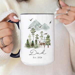 May include: White ceramic mug with a black rim and handle. The mug features a watercolor illustration of a forest scene with mountains, trees, a deer, and birds. The text "Dad Est. 2024" is printed on the mug.