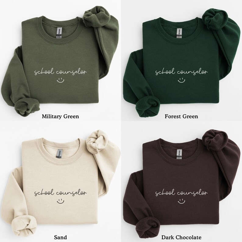 Pode incluir: Quatro sweatshirts de gola redonda em diferentes cores: verde militar, verde floresta, areia e chocolate escuro. Cada sweatshirt tem o texto "school counselor" e um rosto sorridente estampado na frente.