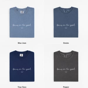 May include: Four folded t-shirts in different colors: blue jean, denim, true navy, and pepper. Each shirt has the phrase "focus on the good" and a smiley face printed on the front. The shirts are displayed on a white background.