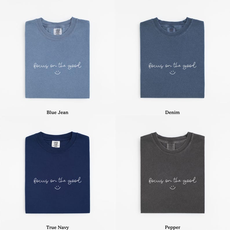 May include: Four folded t-shirts in different colors: blue jean, denim, true navy, and pepper. Each shirt has the phrase "focus on the good" and a smiley face printed on the front. The shirts are displayed on a white background.