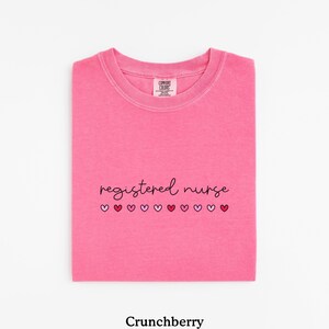 Pode incluir: Uma camiseta rosa brilhante dobrada com as palavras "registered nurse" em fonte cursiva, com uma fileira de cora&ccedil;&otilde;es rosa e vermelhos por baixo. A camiseta est&aacute; rotulada como "Crunchberry" na parte inferior.