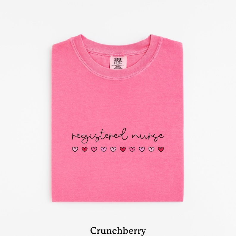Pode incluir: Uma camiseta rosa brilhante dobrada com as palavras "registered nurse" em fonte cursiva, com uma fileira de cora&ccedil;&otilde;es rosa e vermelhos por baixo. A camiseta est&aacute; rotulada como "Crunchberry" na parte inferior.