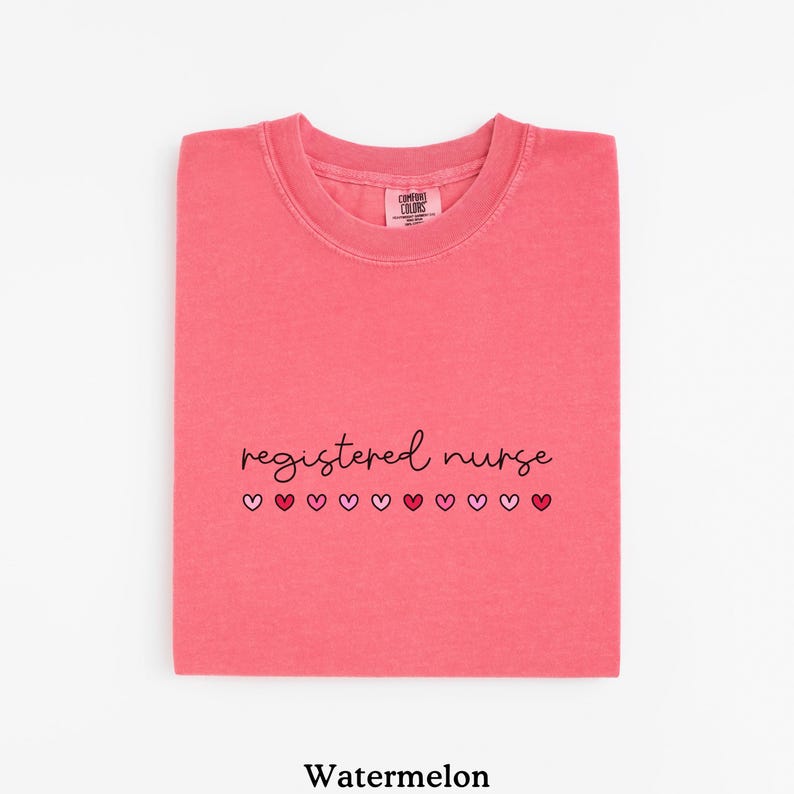 Pode incluir: Uma camiseta dobrada na cor de melancia com as palavras "registered nurse" em escrita preta, seguida por uma fileira de cora&ccedil;&otilde;es rosa e vermelhos. A camisa tem gola redonda e uma etiqueta que diz "Comfort Colors".