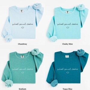 Puede incluir: Cuatro sudaderas con cuello redondo en diferentes colores: Chambray, menta calc&aacute;rea, espuma de mar y azul topacio. Cada sudadera tiene el texto "school social worker" y una carita sonriente impresa en la parte delantera.