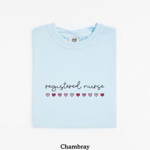 Pode incluir: Camiseta azul clara com a frase "registered nurse" em fonte cursiva, seguida por uma fileira de cora&ccedil;&otilde;es rosa. A palavra "Chambray" est&aacute; impressa na parte inferior da camiseta.