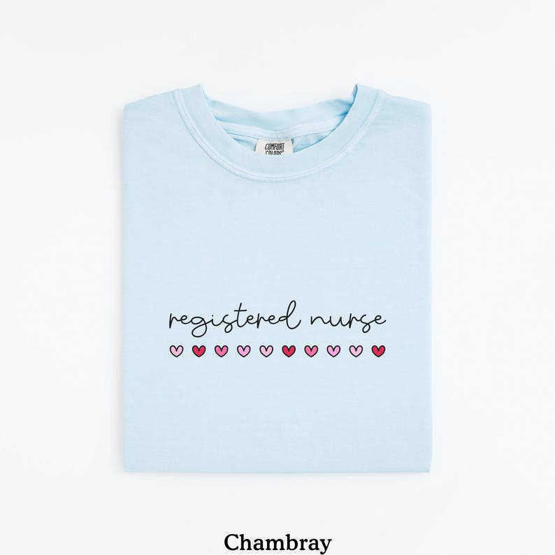 Pode incluir: Camiseta azul clara com a frase "registered nurse" em fonte cursiva, seguida por uma fileira de cora&ccedil;&otilde;es rosa. A palavra "Chambray" est&aacute; impressa na parte inferior da camiseta.