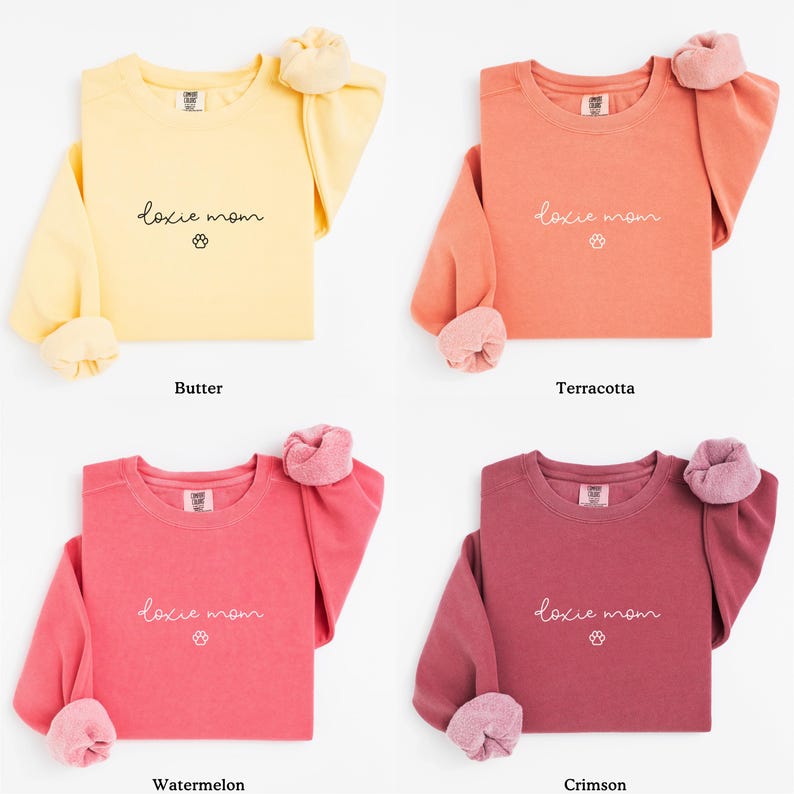 May include: Four long-sleeved sweatshirts in butter yellow, terracotta, watermelon, and crimson. Each sweatshirt has the text "doxie mom" with a paw print. The sleeves are cuffed with a contrasting, fuzzy material.