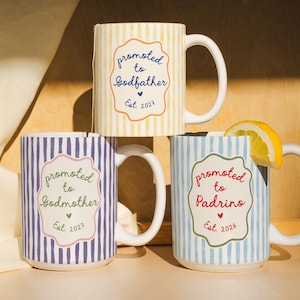 May include: Three ceramic mugs with vertical stripes in yellow, purple, and light blue. Each mug features a scalloped frame containing text: Promoted to Godfather Est. 2027, Promoted to Godmother Est. 2027, and Promoted to Padrino Est. 2026.