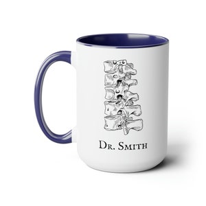 May include: White ceramic coffee mug with a blue rim. The mug features a black and white illustration of a human spine and the text "Dr. Smith".
