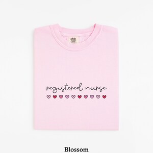 Pode incluir: Camisola rosa claro com a frase "registered nurse" em fonte cursiva e uma fileira de cora&ccedil;&otilde;es rosa por baixo. A camisola &eacute; da marca Comfort Colors.