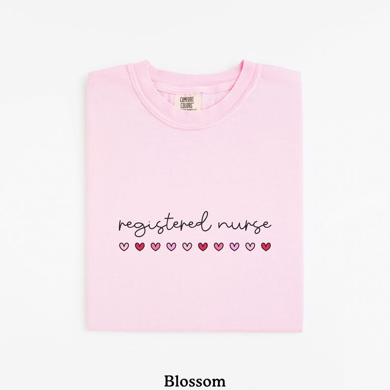 Pode incluir: Camisola rosa claro com a frase "registered nurse" em fonte cursiva e uma fileira de cora&ccedil;&otilde;es rosa por baixo. A camisola &eacute; da marca Comfort Colors.