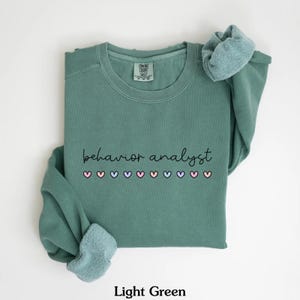 K&ouml;nnte beinhalten: Hellgr&uuml;ner Pullover mit dem Text "behavior analyst" in schwarzer Schrift und einer Reihe von rosa, blauen und violetten Herzen.