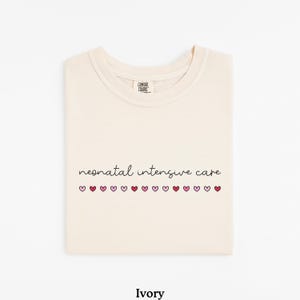 May include: Ivory t-shirt with the text "neonatal intensive care" and a row of pink and red hearts.