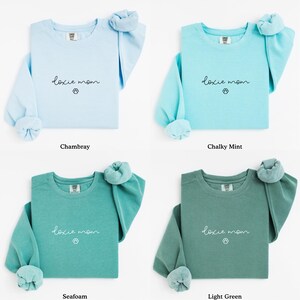 May include: Four long-sleeved sweatshirts in chambray, chalky mint, seafoam, and light green. Each shirt features the text "doxie mom" with a paw print graphic. The sleeves are rolled up, and the shirts are displayed on a white background.